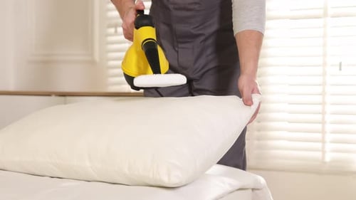 Man Steaming White Pillow with Yellow Steamer in Bedroom