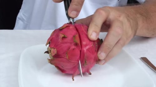 A person is using a kitchen knife to slice a vibrant pink dragon fruit on a plate placed on the tabl