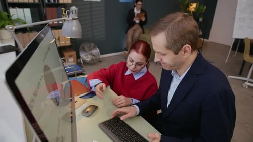Young Woman Assisting Adult Colleague Doing Diagram on Computer in Office