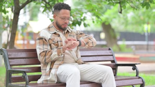 Hispanic Man Using Smart Watch on Park Bench