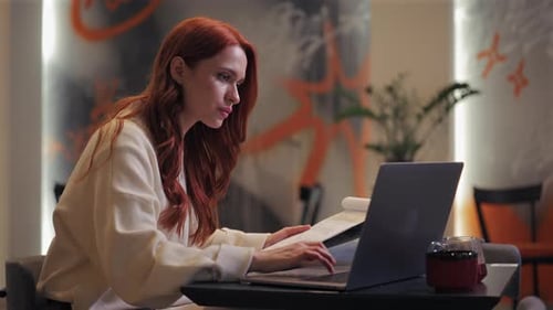 Side View of Redhaired Caucasian Woman Studying Documents While Sitting at Table with Laptop Female
