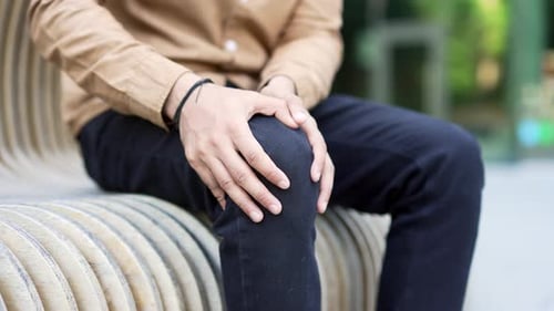 Male hands massaging knee leg muscles while sitting on a bench on street near an office building.