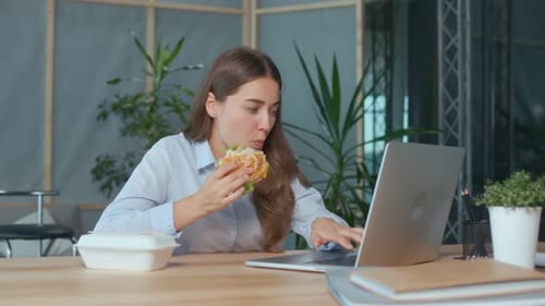 Junge Geschäftsfrau isst Hamburger, während sie am Laptop am Bürotisch arbeitet