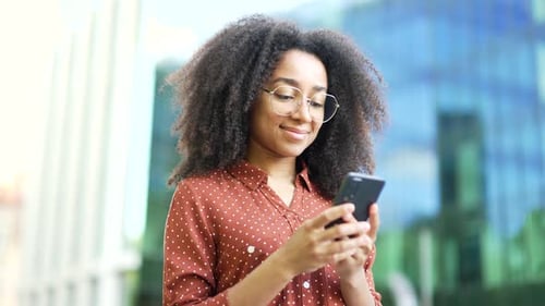 African American woman using smartphone outdoors in urban area smiling. Person holding phone wearing