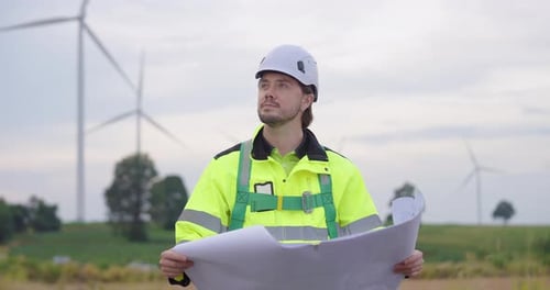 Male engineer wearing safety helmet reading blueprint at wind farm, concentrating on renewable