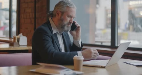 Male Entrepreneur Working with Laptop and Discussing Business on Mobile Phone Indoors in Cafe