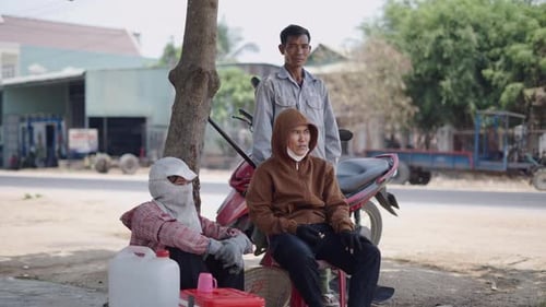 Three Asian People Rest and Chat Quietly Under a Tree Near the Road in Vietnam