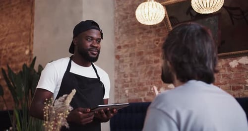 A Black Person in a Black Apron and Cap Takes an Order From a Customer Using an Electronic Tablet