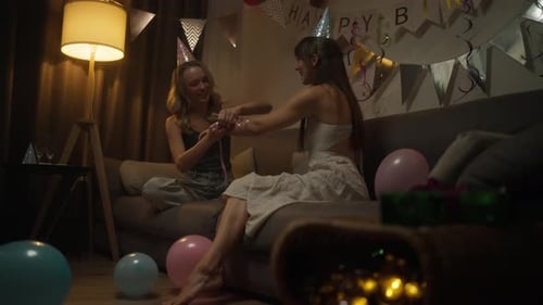 Happy Brunette Girl in a Pink Party Cap Decorates Her Friends Hand with a Pink Ribbon While Having