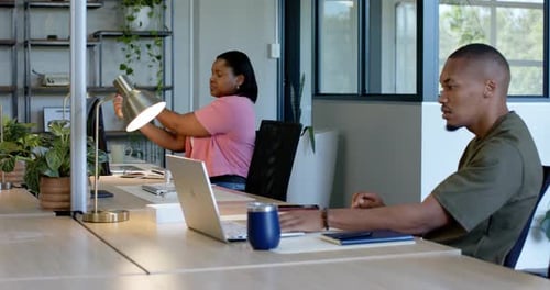 Collègues africaines au bureau, femme qui s'étire, ajuste la lampe pour la lumière pendant qu'un homme tape sur un ordinateur portable