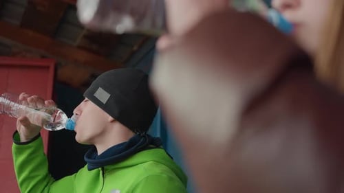 Two Players Drink Water on Bench Caucasian Male in Green Jacket with Teammate Blurred Bottles Raised