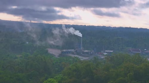 Aerial view of factory chimney emitting smoke from combustion. Factory surrounded by green trees.