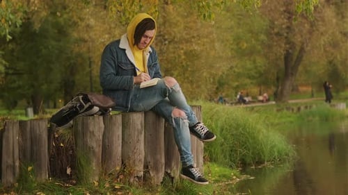 Young Creative Man with Dreadlocks Drawing Writing in Notebook Sitting on Stumps Near Lake in