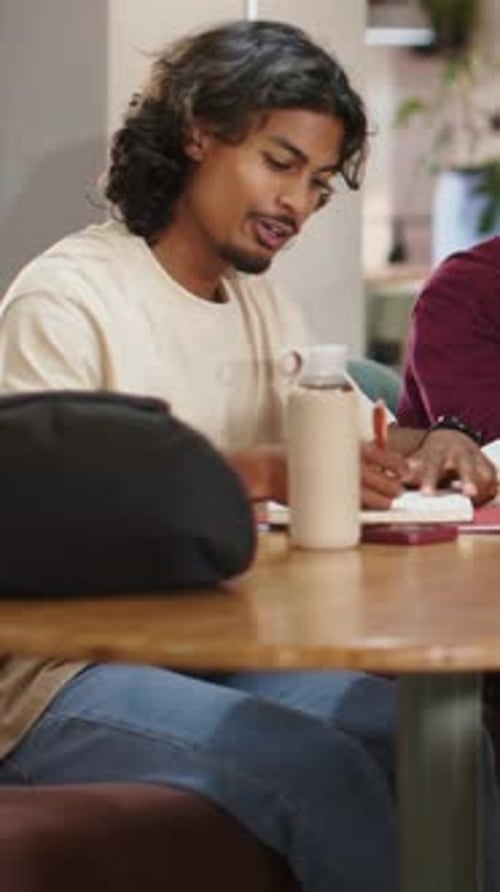 Vertical video: Writing student in beige tee, starting notes, pointing notebook, discussing at cafe