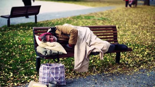 Homeless man sleeps rough on park bench with pillow in city street motion