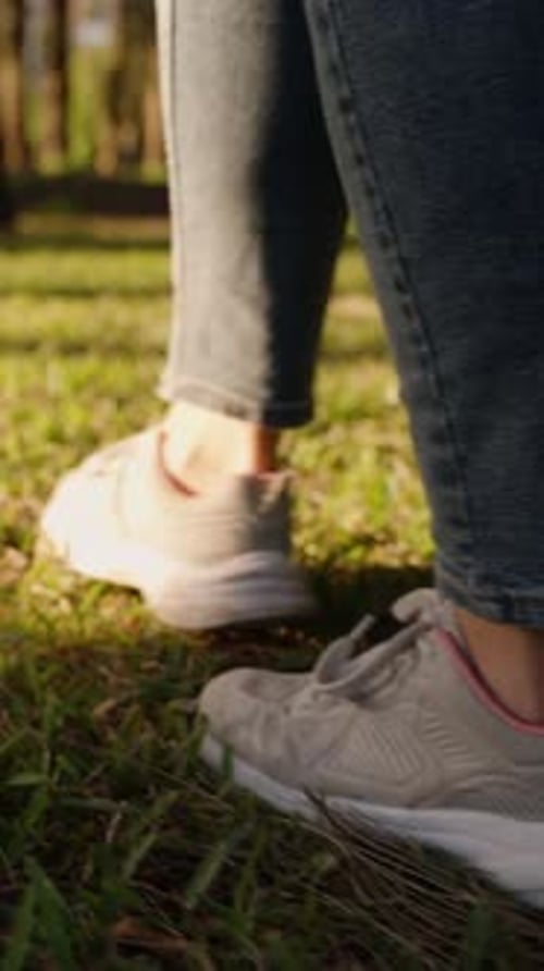 Vertical video. Slow Motion. Stroll through the Forest at Sunset, Female Legs in Sneakers on Green G