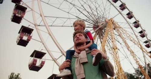 A Little Blond Boy with Blue Eyes in a Red Tshirt Sits on the Shoulders of His Dad with Curly Hair