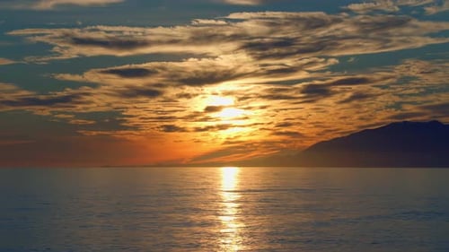 Dramatic sunset with calm ocean waves, clouds, and mountains on the background