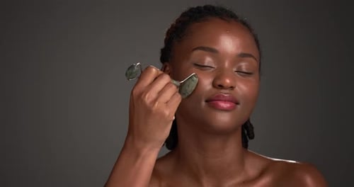 Happy, black woman or skincare with jade roller in studio for anti aging on a gray background
