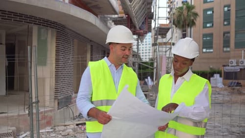 Two diverse Architects in safety vests and helmets walking and checking blueprints outdoors