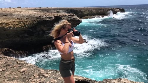 Beautiful blonde woman taking picture on a cliff edge with wavy blue ocean background on a sunny day