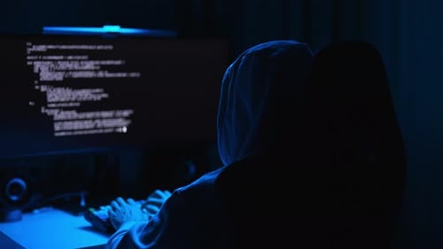 Anonymous Male Hacker Wearing Sweatshirt with Hood Typing on Keyboard Looking to Monitor with Text