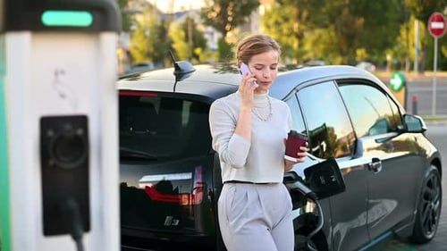 A young blonde woman with coffee talking on the smartphone at a car charging station with electric c