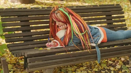 Young Woman with Dreadlocks Drawing Writing in Notepad Lying on Park Bench in Golden Autumn