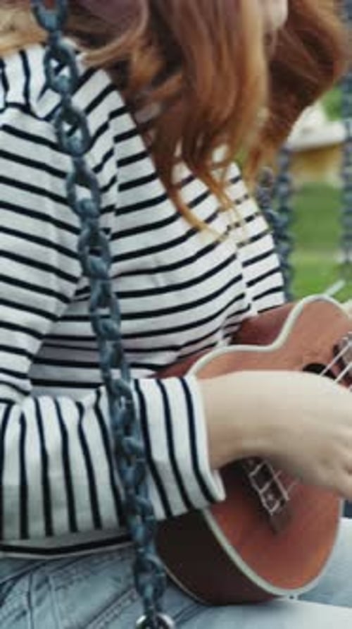 Young Woman Practices Playing The Ukulele Seated In A Swing