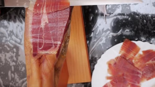 Overhead, closeup shot, Spanish cured ham being sliced with long thin knife. Meat is dark red colour