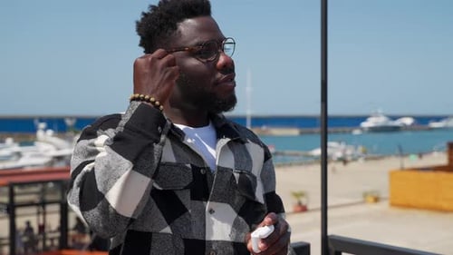 Black Man is Putting Inear Earphones and Enjoying Music From Smartphone Portrait in City Near Sea