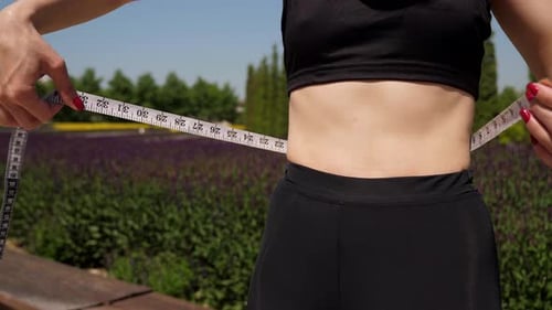 A Closeup of a Slender Young Woman Measures Her Waist in a Sunny City Park