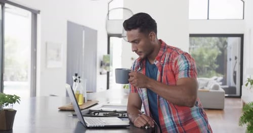 Biracial man having coffee and using laptop in sunny modern apartment, slow motion