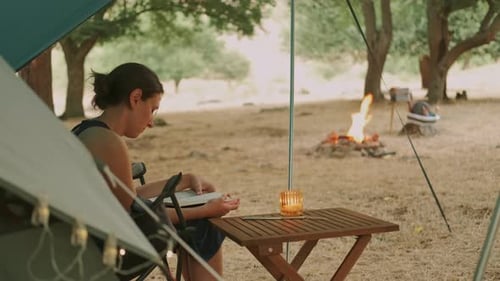 Woman Reads by Tent and Campfire in Nature