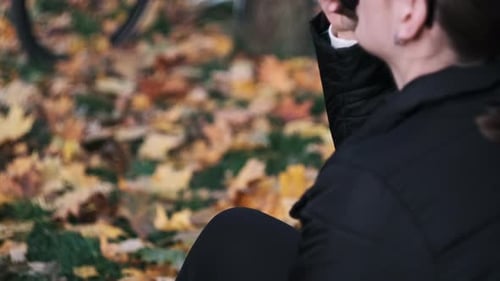 Young Woman Drinks Coffee on Lawn in Autumn Park Among Fallen Yellow Leaves