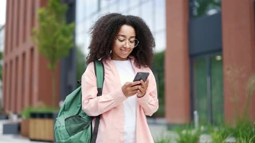 Smiling african american female student using phone standing on campus space near university