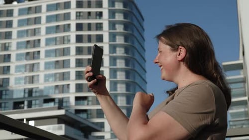 Happy Caucasian Woman Takes Pictures of Modern City From Balcony on Phone