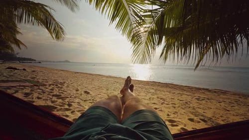 First Person View Of Legs Stretching In Hammock Toward Tropical Beach With Palm Leaves And Sparkling