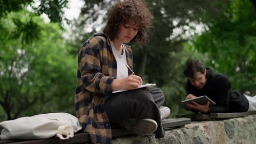 A Brunette Student Girl with Curly Hair in a Brown Shirt Sits on a Bench and Makes Notes in a