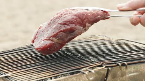 I place a juicy beef steak on my portable grill using tongs, with the beach as the backdrop.