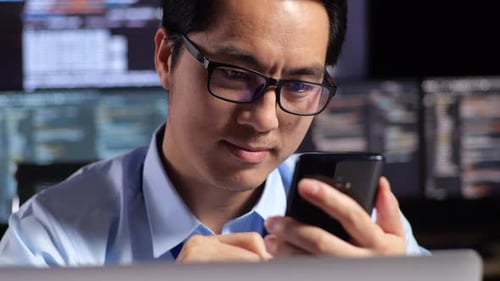 Primer plano del programador que usa un teléfono inteligente mientras escribe código con una computadora portátil usando varios monitores