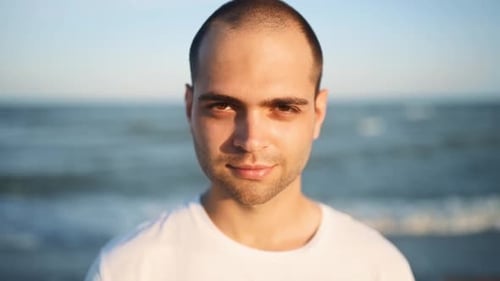 Portrait of Young Handsome Man Looking and Smiling at Camera Standing on the Beach with a Sea on