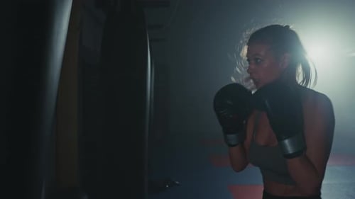 Focused Woman Trains Intensely on a Punching Bag During Evening Workout Session in a Dimly Lit Gym