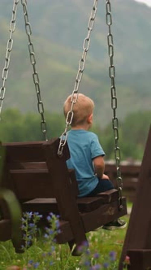 Lonely Boy Sits on Swings on Playground Against Mountains
