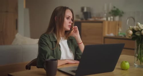 A Girl in a Green Jacket Talks on the Phone While Working on a Laptop During the Day at Home