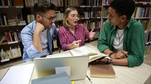 Three young students study in the school library, talking and using laptop for researching online.