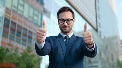 A man in a suit giving two thumbs ups with a big smile on his face standing outside Glass Skyscraper