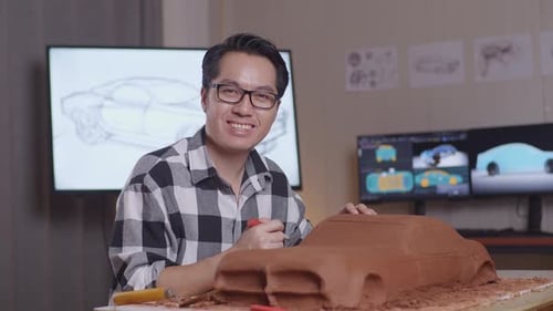 Asian Man Designer Smiling To The Camera While Working On The Sculpture Of Car Clay In The Studio