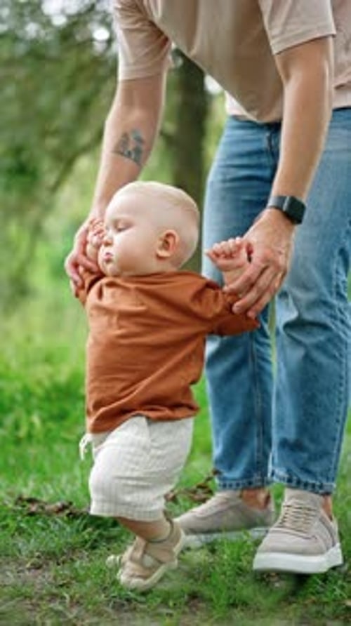 Blond toddler wearing brown t-shirt and white shorts walks supported by dad.