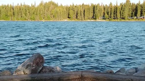 Zooming in on clear, blue lake surrounded by lush green vegetation and cloudy blue sky. Water is sli